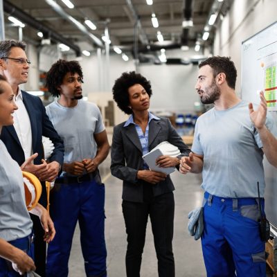 Young manual worker presenting new business strategy to company managers and his colleagues in a factory.